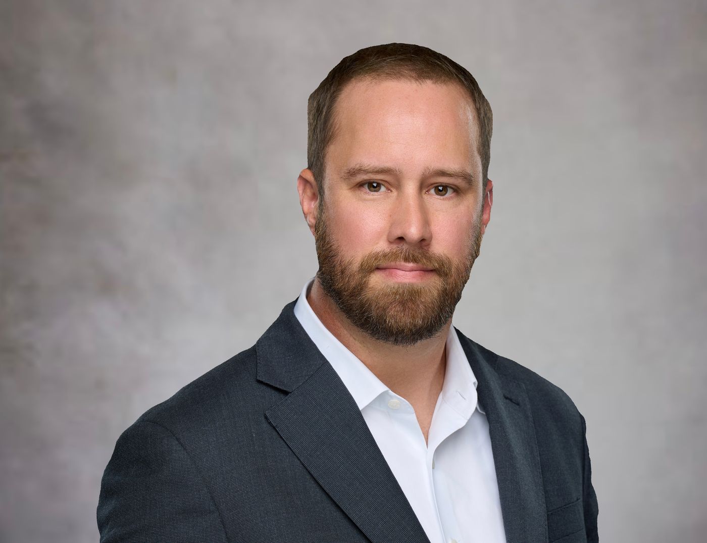 Ken Dickinson, wearing a charcoal blazer and open-collar white shirt, photographed against a plain gray studio backdrop.
